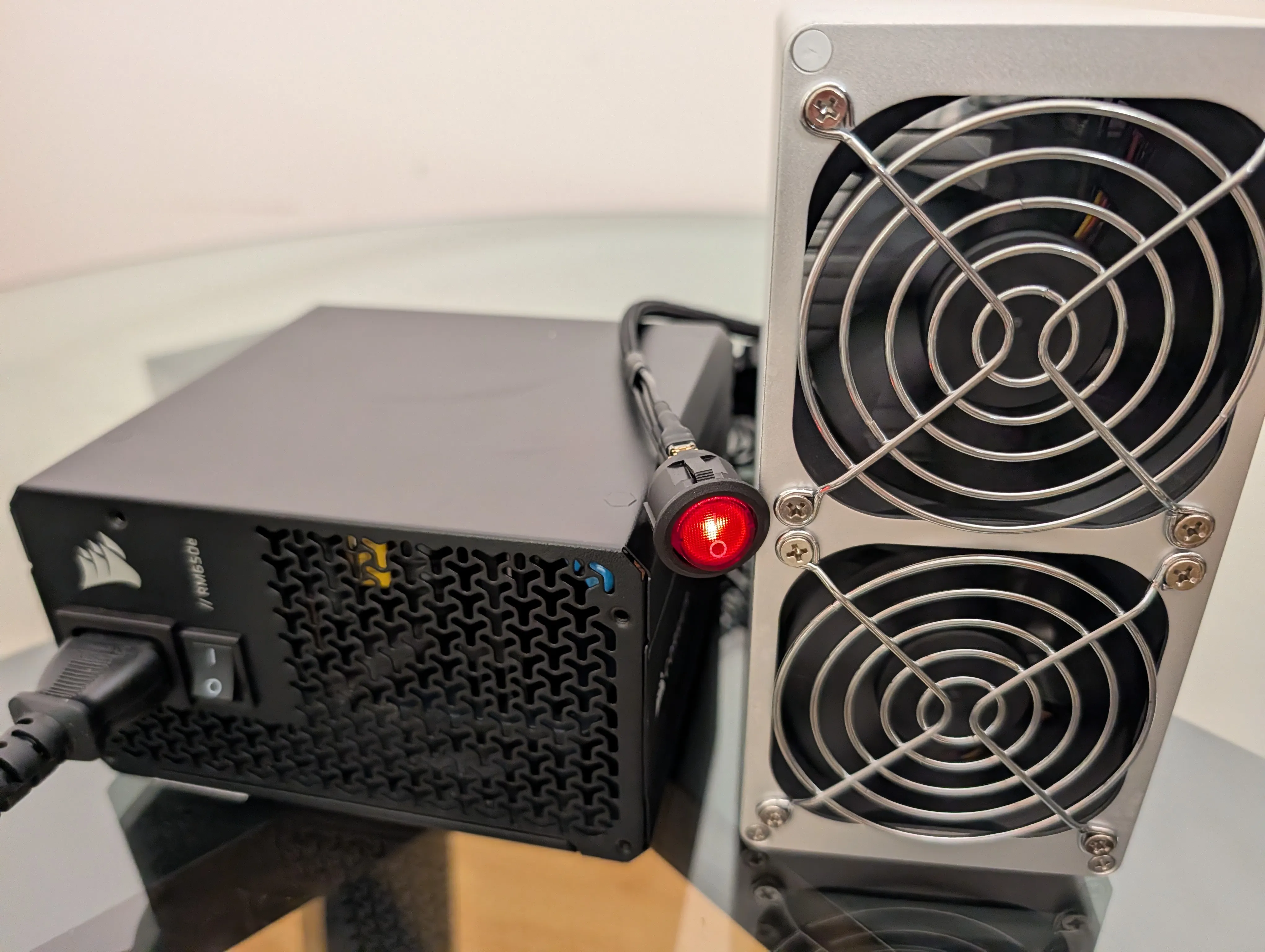 The Corsair RM650e power supply with its red rocker jumper switch lit, sitting next to the silver Goldshell SC Box with its two fan grilles, on a glass table